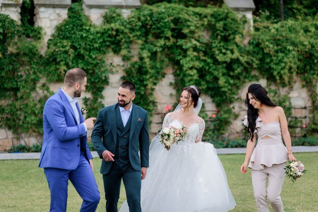 Pareja de novios con mejores amigos está sonriendo al aire libre cerca del muro de piedra cubierto de hiedra