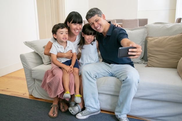 Pareja de padres alegres y dos niños sentados en el sofá en casa juntos, tomando selfie