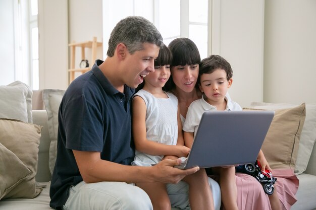 Pareja de padres alegres emocionados con niños en el regazo, sentados en el sofá todos juntos, viendo películas o videos en la computadora portátil en casa, mirando la pantalla.