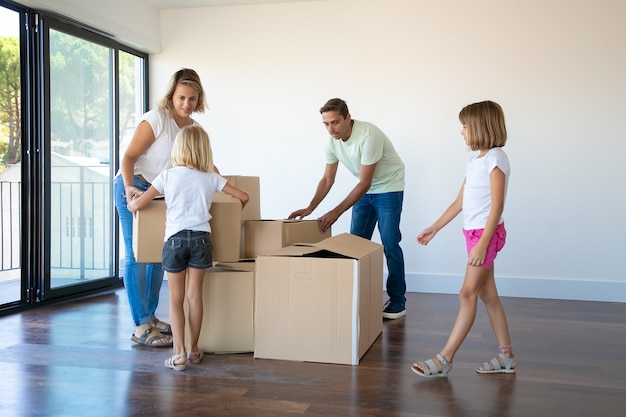 Pareja de padres y dos niñas abriendo cajas y desempacando cosas en su nuevo piso vacío