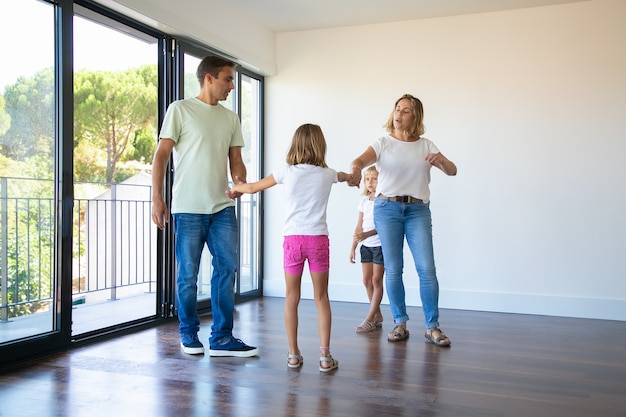 Pareja de padres y dos niños disfrutando de su nuevo hogar, de pie en una habitación vacía y tomados de la mano, bailando