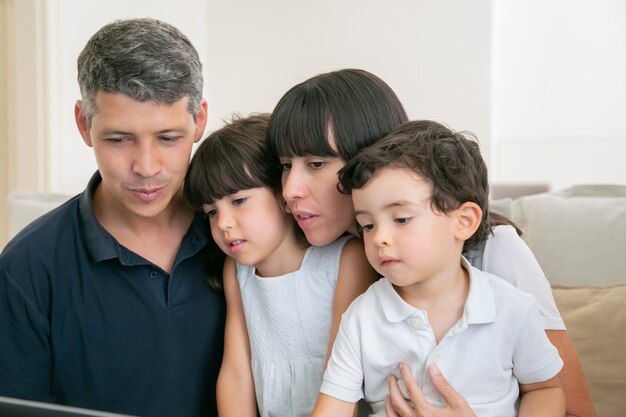 Pareja de padres y dos niños usando la computadora para videollamadas, sentados juntos en el sofá, mirando la pantalla