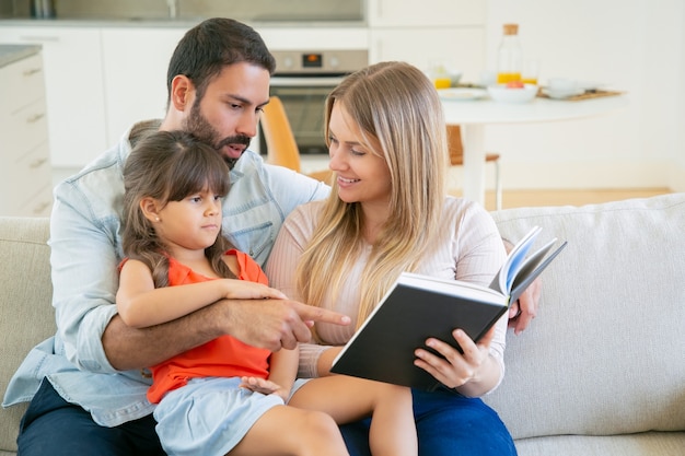 Pareja de padres felices y niña de pelo negro sentada en el sofá en la sala de estar y leyendo un libro juntos.