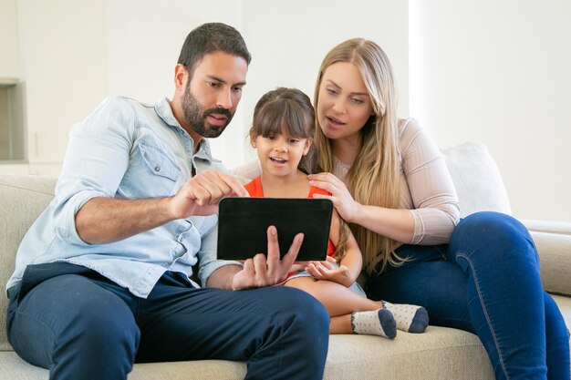 Pareja de padres jóvenes y linda hija sentada en el sofá, usando tableta para videollamadas o ver películas.