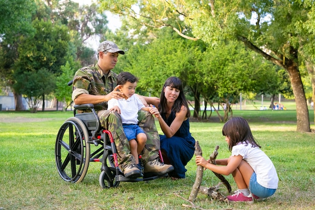 Pareja de padres pensativos y pacíficos que pasan tiempo libre con los niños al aire libre, preparando leña para fuego en el césped Veterano discapacitado o concepto de familia al aire libre