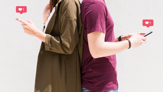 Pareja de pie espalda con espalda usando el teléfono móvil con los iconos de mensajes de amor contra la pared