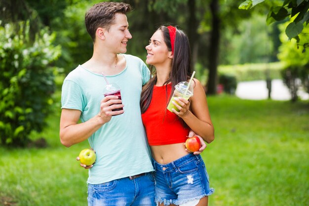 Pareja de pie en el parque con batidos saludables y manzanas mirando el uno al otro