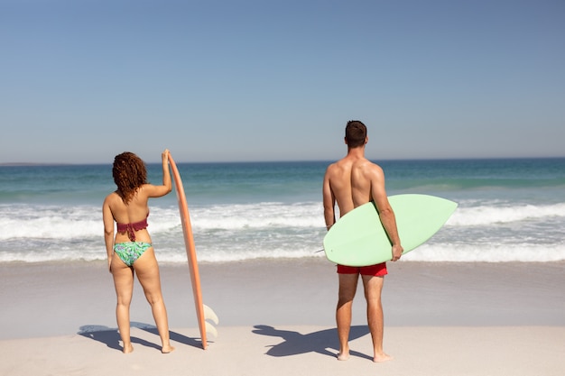 Pareja de pie con tabla de surf en la playa bajo el sol