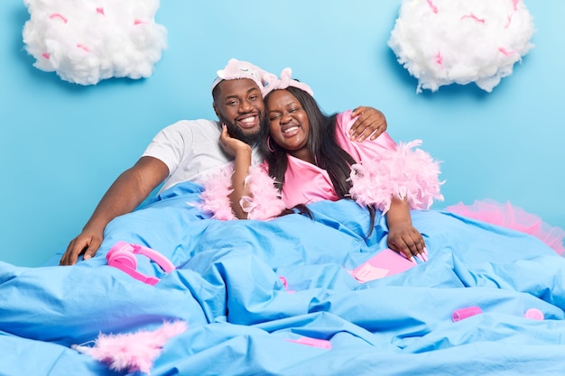 Pareja de piel oscura positiva abrazarse en la cama, amarse mutuamente, disfrutar de pasar tiempo juntos en casa, tener una sonrisa de día perezoso ampliamente aislado en azul