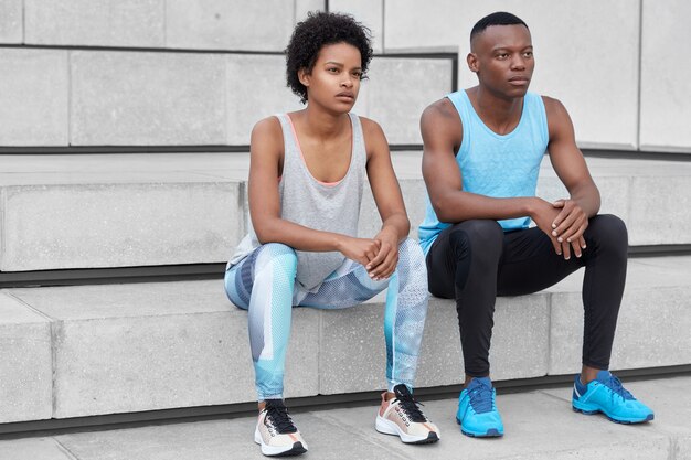 Pareja de piel oscura vestida con ropa deportiva, zapatillas cómodas, tomar un descanso después del entrenamiento cardiovascular, sentarse en las escaleras de la ciudad, concentrarse en la distancia, estar un poco cansado. Novio y novio aficionado al deporte