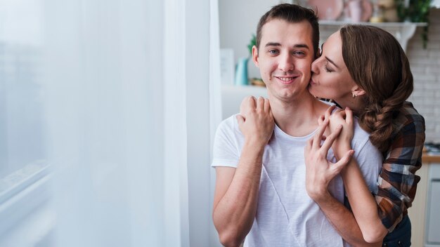 Pareja positiva abrazando y besando cerca de la ventana