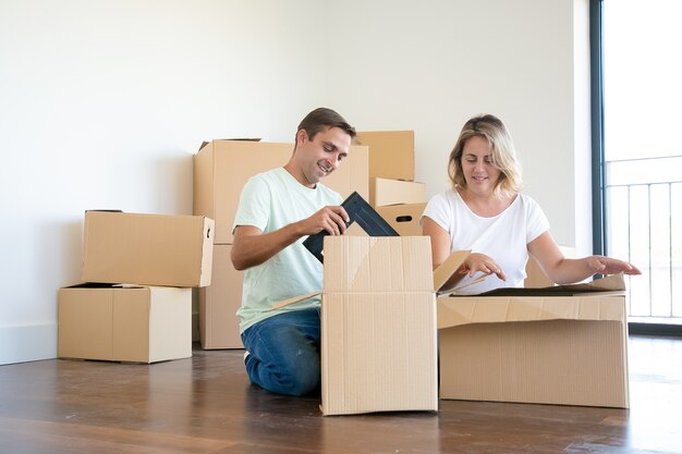 Pareja positiva desembalaje de cosas en un apartamento nuevo, sentado en el suelo y tomando objetos de cajas abiertas