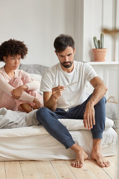 Una pareja de raza mixta desesperada se entera de un embarazo no deseado, mira con expresión triste en la prueba, posa juntos en el dormitorio en casa, se siente decepcionado. La anticoncepción falla y el concepto de fertilidad.