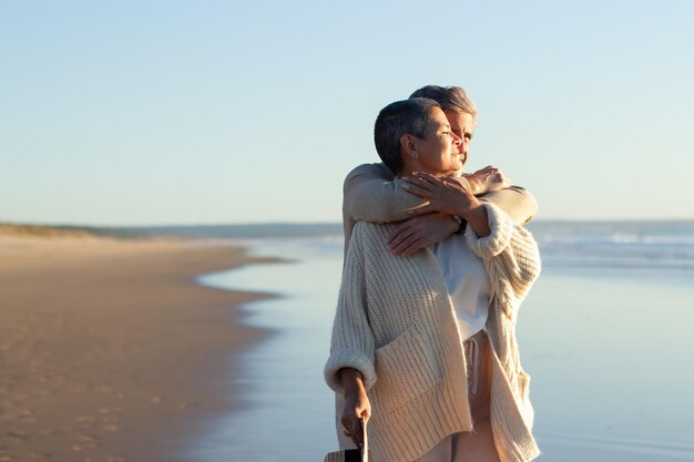 Una pareja romántica de ancianos pasa tiempo en la orilla del mar al atardecer, disfrutando de fantásticas vistas al mar. Hombre canoso abrazando a su esposa mientras está de pie detrás. Romance, jubilación, concepto de vacaciones.