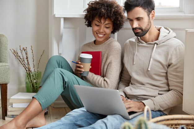 Una pareja romántica se sienta cerca en el piso, enfocada en la computadora portátil, mira películas interesantes en línea, disfruta de un café aromático, está de buen humor, disfruta de la conexión inalámbrica a Internet, tiene tiempo libre