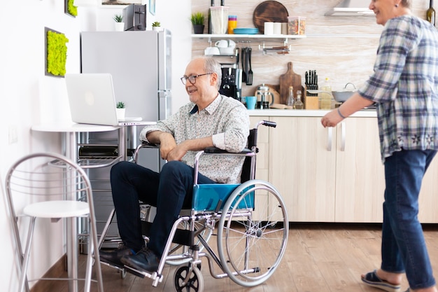 Pareja senior riendo usando laptop, esposo llamando a su esposa cerca de él durante una videollamada con nietos sentados en la cocina. Anciano discapacitado paralizado usando tecnología de comunicación