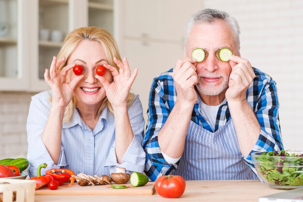 Pareja senior sonriente sosteniendo tomates cherry y rodajas de pepino frente a sus ojos