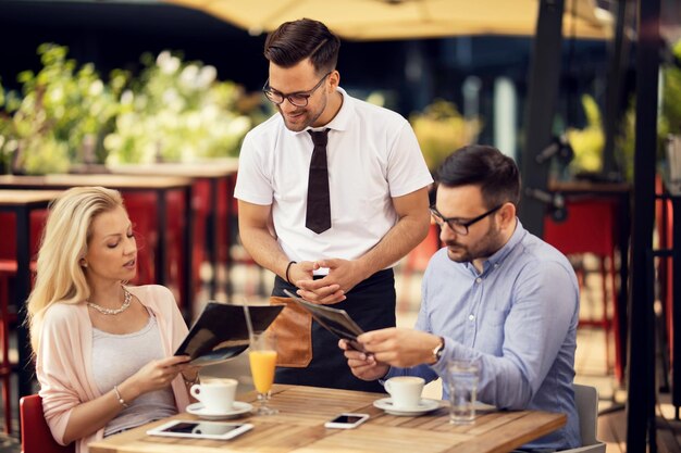 Pareja sentada en un café eligiendo el pedido de un meu mientras se comunica con un camarero