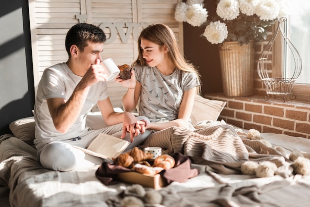 Pareja sentada en la cama cogidos de la mano del otro desayunando juntos