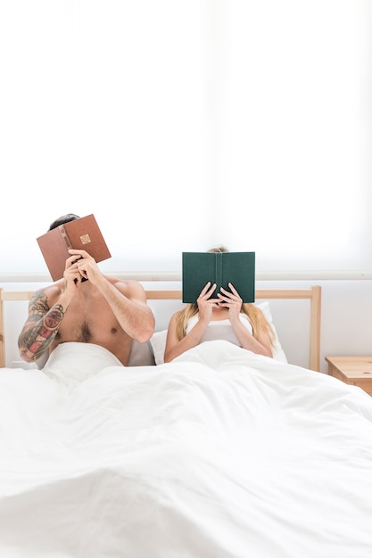 Pareja sentada en la cama escondiendo su cara con el libro