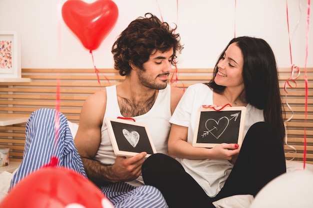 Pareja sentada en la cama con pizarras negras con corazones dibujados