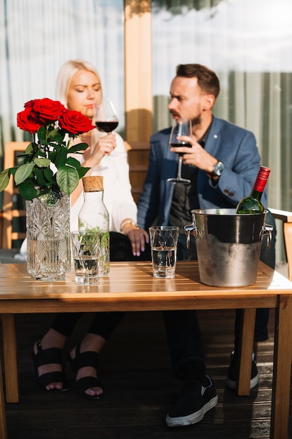 Pareja sentada delante de la botella de vino en un cubo de hielo en la mesa con rosas