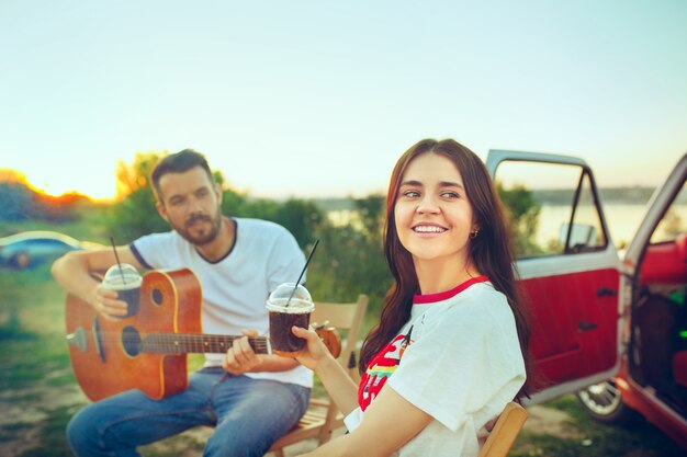 Pareja sentada y descansando en la playa tocando la guitarra en un día de verano cerca del río. Amor, familia feliz, vacaciones, viajes, concepto de verano.