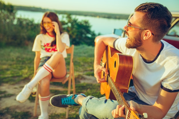 Pareja sentada y descansando en la playa tocando la guitarra en un día de verano cerca del río.