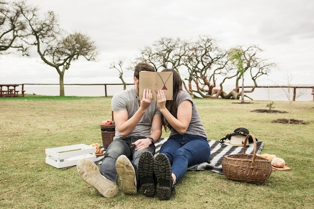 Pareja sentada en la manta sosteniendo el libro sobre su cara en picnic