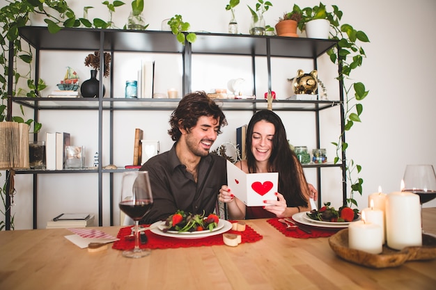 Pareja sentada en una mesa para comer mientras ella lee una postal romántica y sonrie