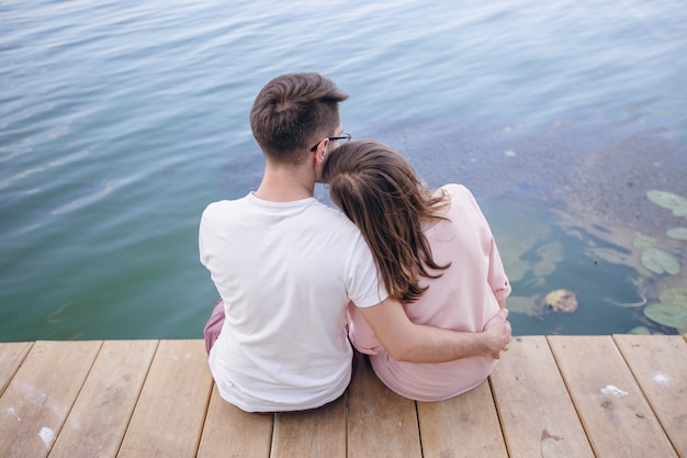 Pareja sentada en un muelle de madera y ella con la cabeza apoyada en el hombro de él