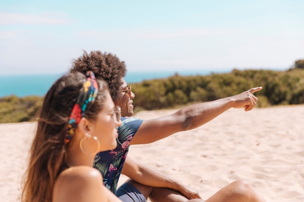 Foto gratuita pareja sentada en la playa y chico mostrando algo lejos a chica