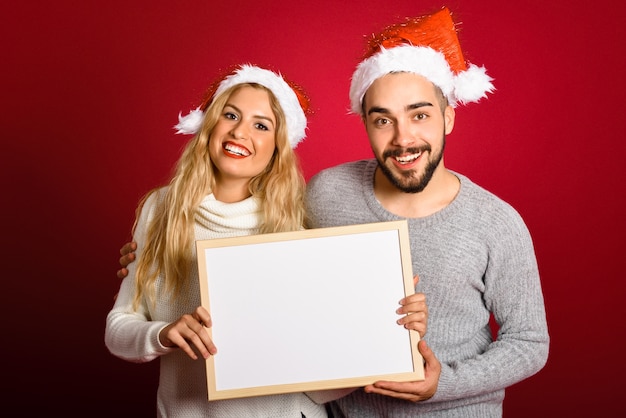 Pareja con un sombrero de de santa sosteniendo un tablero blanco sobre fondo rojo