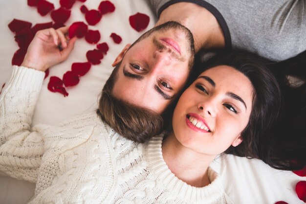 Pareja sonriendo y tumbada en la cama cabeza con cabeza rodeada de pétalos de rosa