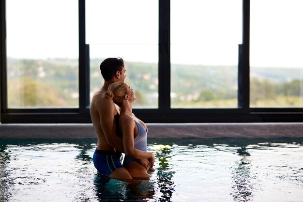 Pareja sonriente abrazándose mientras está de pie en la piscina termal y disfrutando de la vista a través de la ventana en el spa de salud Espacio de copia