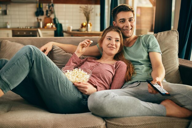 Pareja sonriente comiendo palomitas de maíz y viendo la televisión mientras se relaja en el sofá
