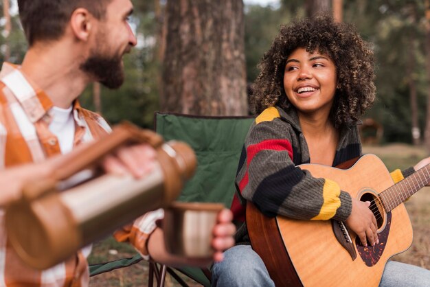 Pareja sonriente disfrutando de acampar al aire libre con guitarra y bebida caliente