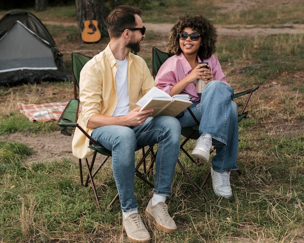 Foto gratuita pareja sonriente con gafas de sol leyendo y bebiendo mientras acampa al aire libre