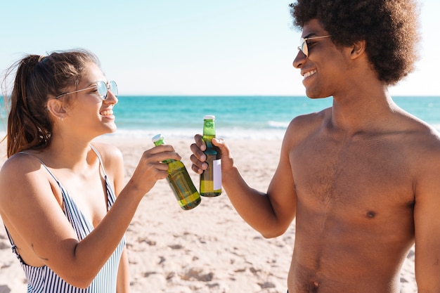 Pareja sonriente de pie en la orilla del mar y tintineo botella de bebida