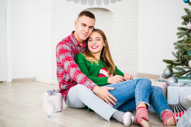 Pareja sonriente sentada al lado de árbol de navidad