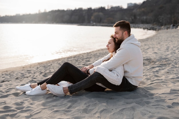 Pareja sonriente sentada en la playa tiro completo