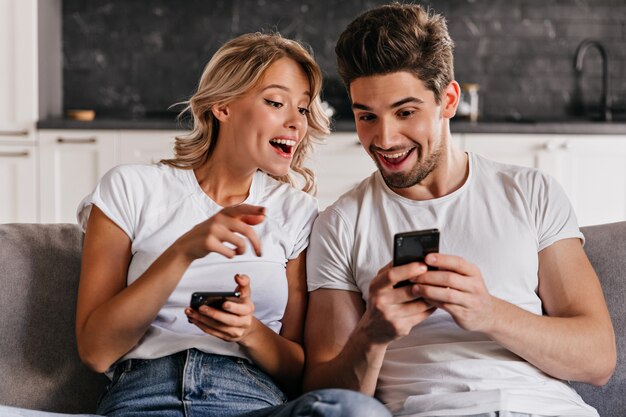 Pareja sonriente sentada en el sofá con teléfonos. Adorable mujer joven con smartphone.