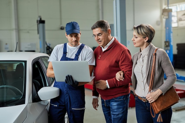 Pareja sonriente y su reparador de automóviles usando una computadora portátil en un taller