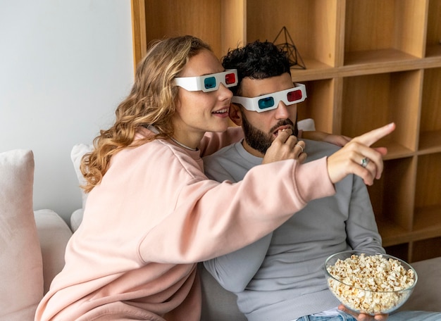 Pareja sonriente viendo películas en casa con gafas tridimensionales y comiendo palomitas de maíz