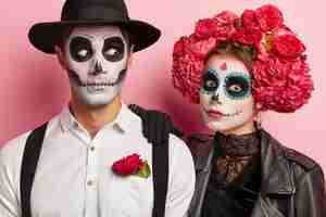 Foto gratuita la pareja sorprendida tiene expresiones faciales aterradoras, maquillaje y disfraces originales, usa atuendo blanco y negro decorado con flores rojas, posa juntos en el estudio contra la pared rosa