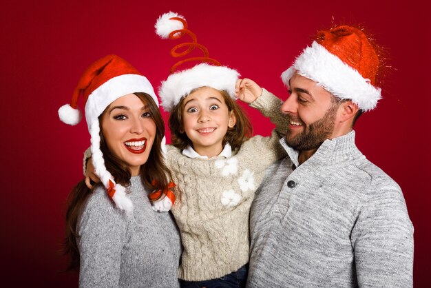 Pareja con su pequeña hija vestida con ropa de invierno y sombrero de santa sobre fondo rojo