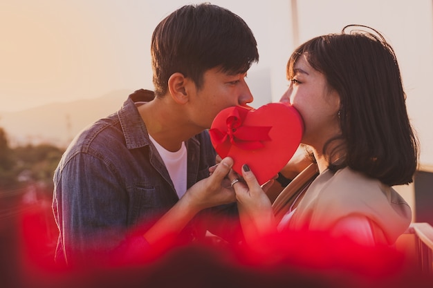 Pareja con tapando sus bocas con una caja de regalo con forma de corazón