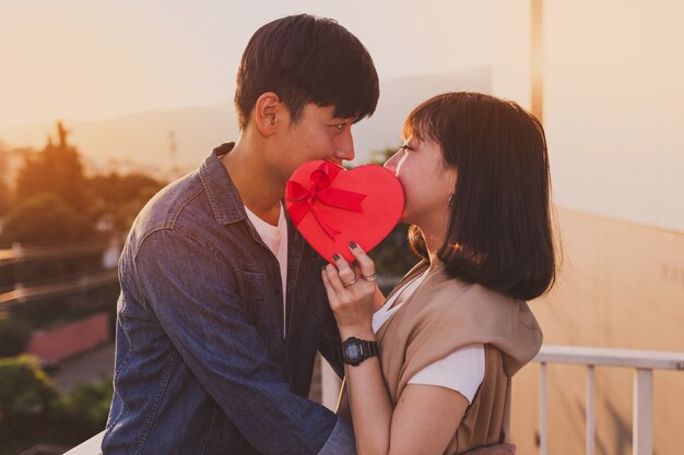 Pareja con tapando sus bocas con una caja de regalo con forma de corazón