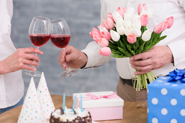 Pareja tintineo de copas de vino con ramo de flores de tulipán; pastel de cumpleaños y cajas de regalo en mesa