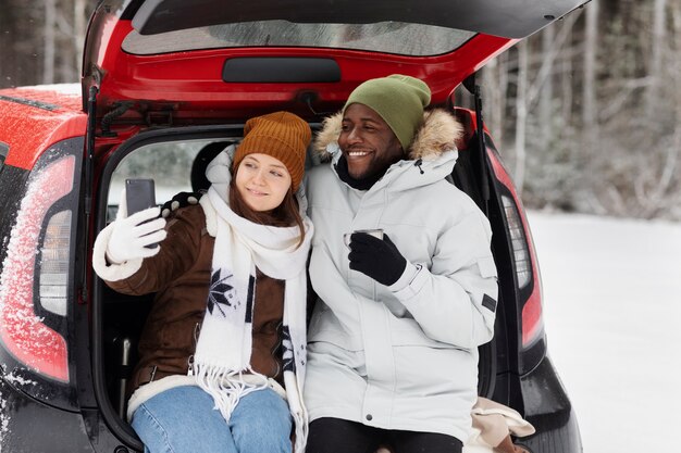 Pareja tomando selfie en el maletero del coche durante un viaje por carretera en invierno